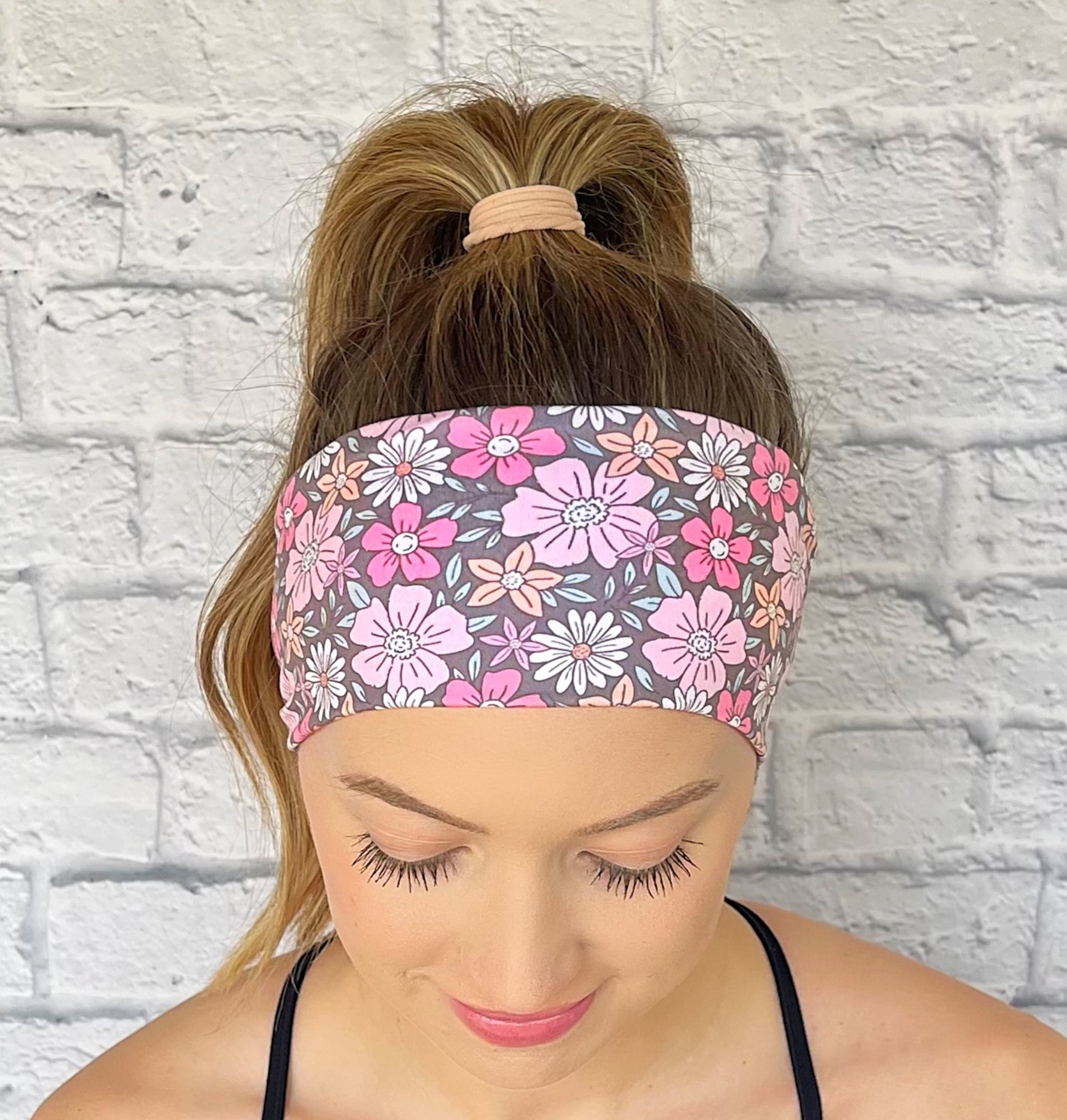 Woman with hair in curled ponytail wearing flat, stretchy, gray headband with pink, peach, and white floral print.