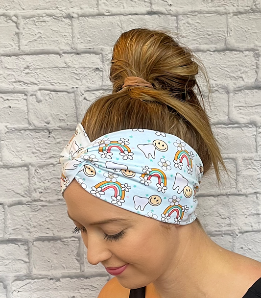 Woman with hair in bun wearing wide, twisted headband in light blue with teeth, rainbows, flowers, and smiley faces print.