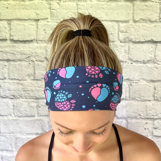 Woman with hair in ponytail wearing stretchy, navy color headband with pink and blue baby feet print.