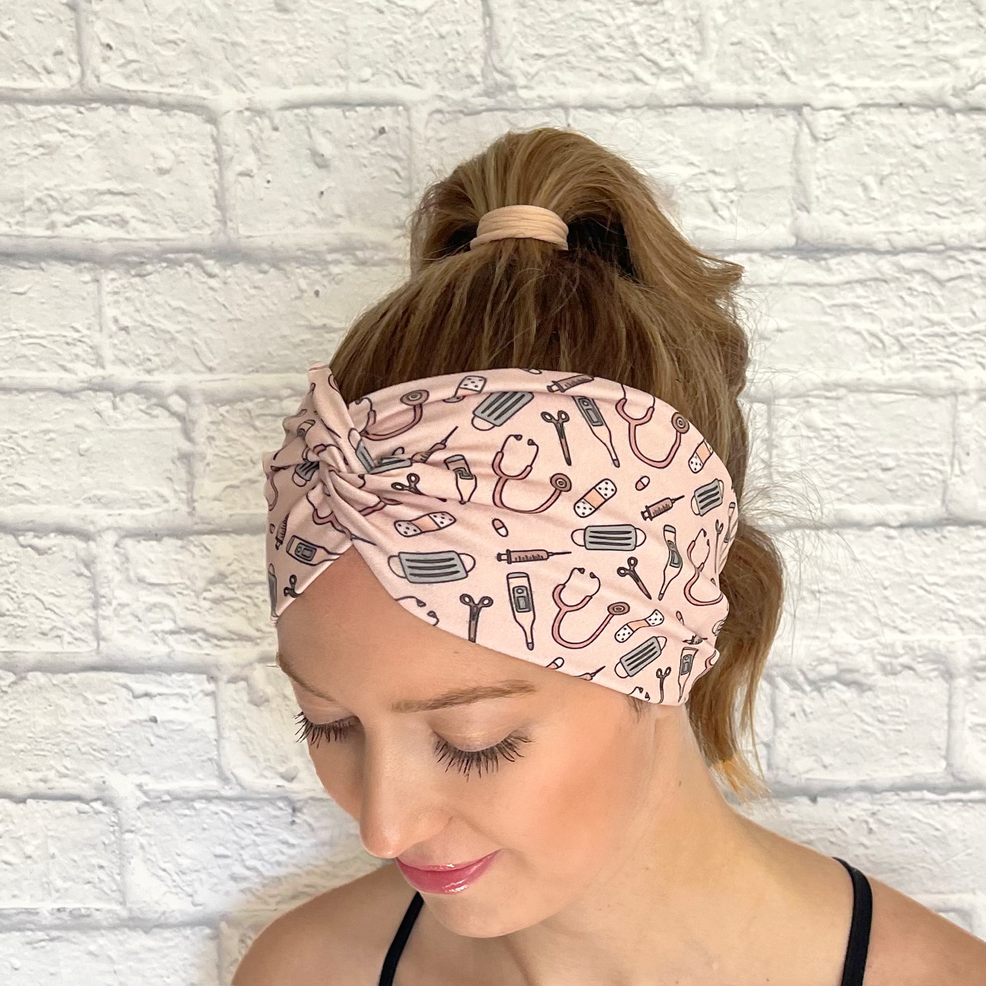 Woman with hair in curled ponytail wearing wide, twisted headband in peach color with medical theme print.