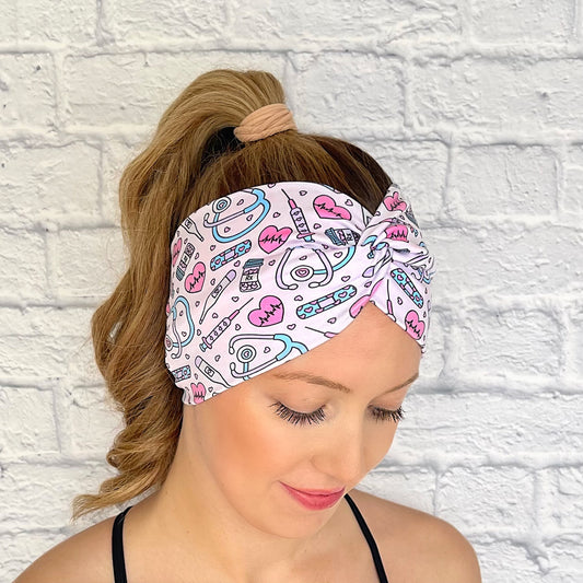 Woman with hair in curled ponytail wearing wide, twisted headband in light pink with pink, purple, and blue medical theme print.