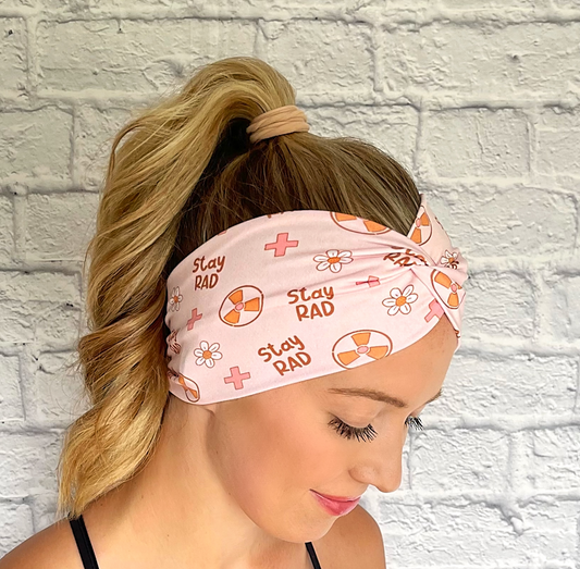 Woman with hair in curled ponytail wearing, twisted, wide headband in pink with "stay rad" print, radiology theme print, and flowers.