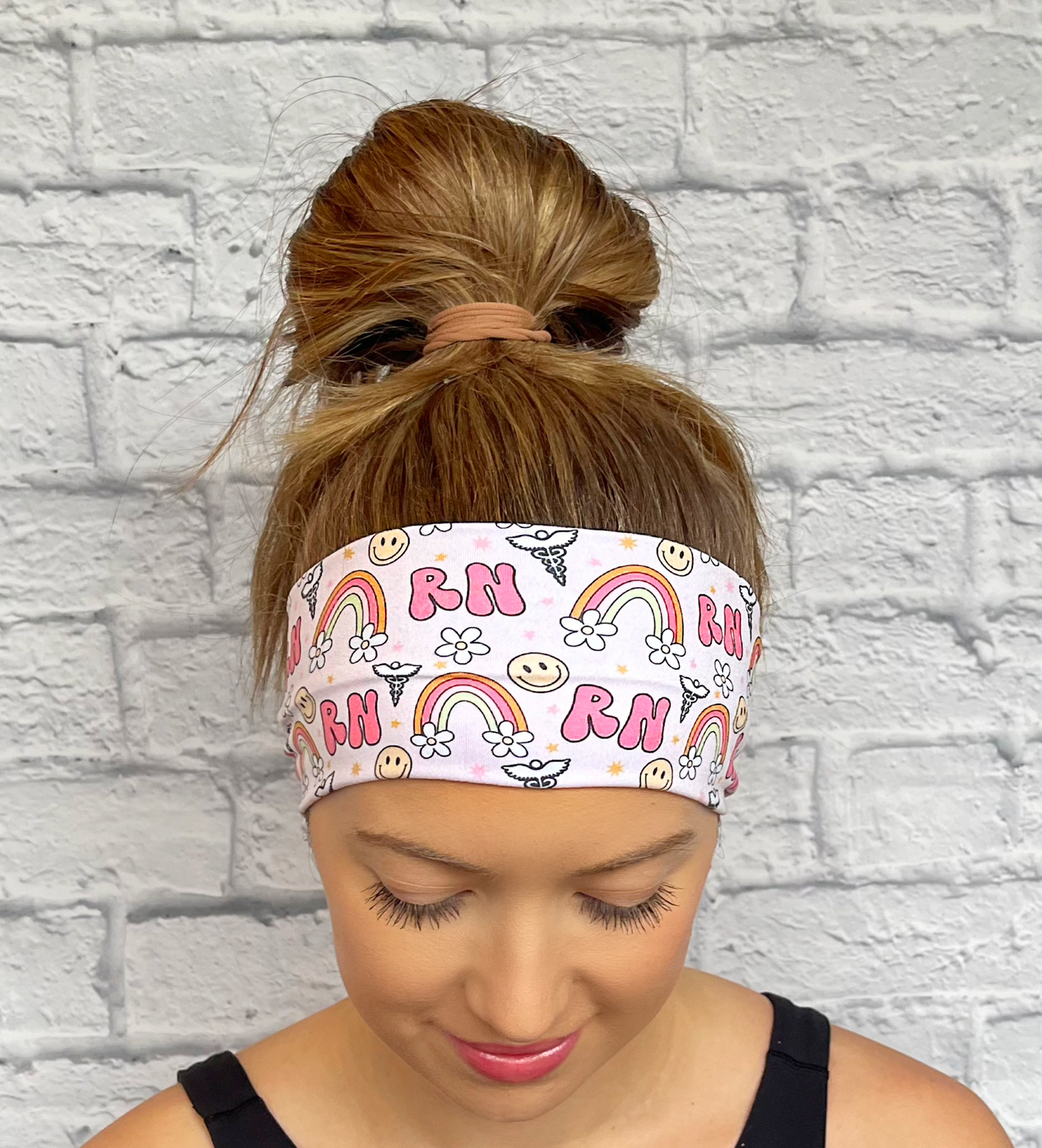 Woman with hair in bun wearing stretchy, light pink headband with "RN", rainbows, flowers, and smiley faces