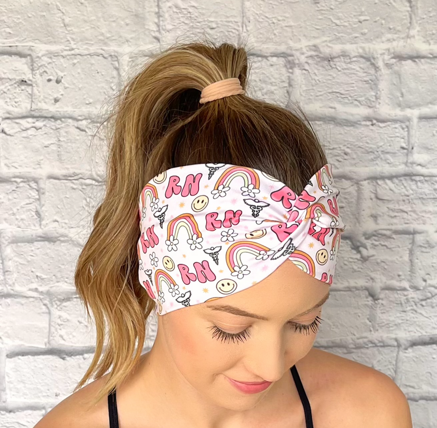 Woman with hair in curled ponytail wearing wide, twisted headband with pink "RN" print, rainbows, flowers, smiley faces, and medical symbol.