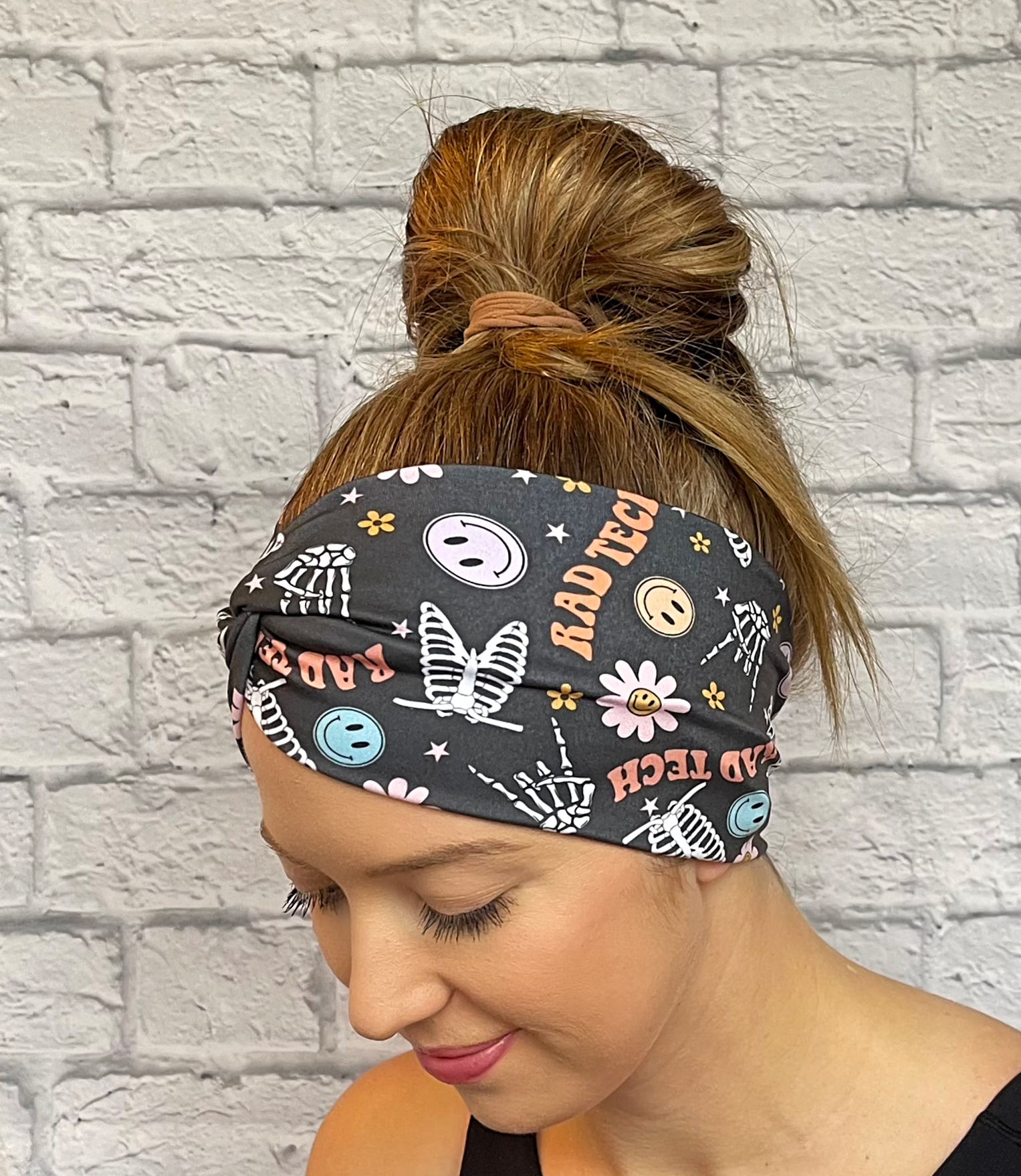 Woman with hair in bun wearing a dark gray, wide, twisted headband with skeletons, flowers, smiley faces, and "rad tech" print.