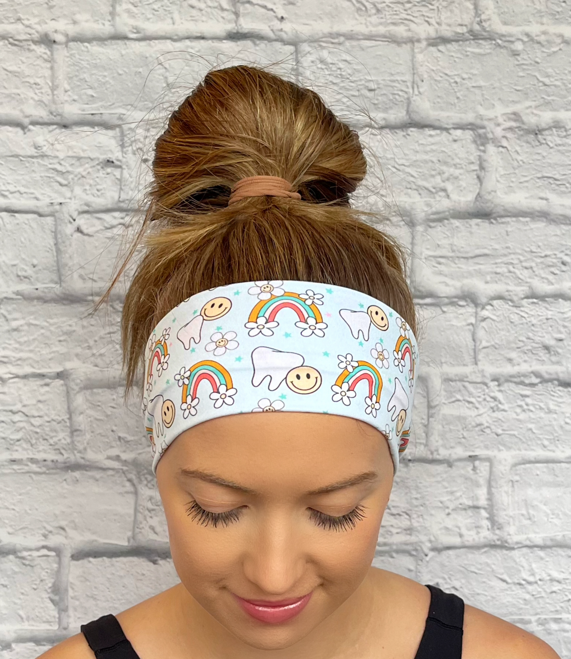 Woman with hair in bun wearing light blue headband with teeth, rainbows, smiley faces, and flowers.