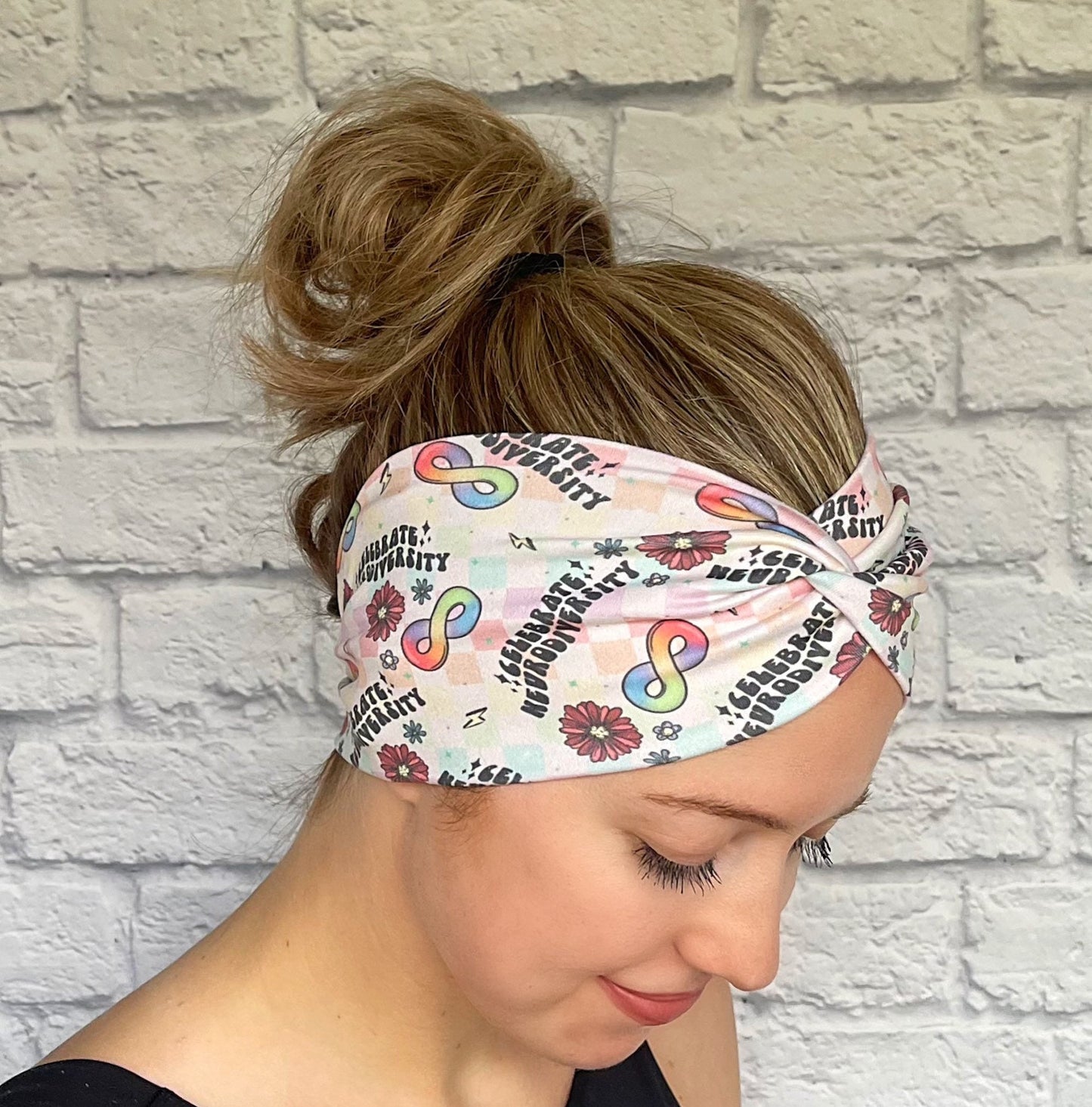 Woman with hair in bun wearing wide, twisted headband in rainbow plaid print with "celebrate neurodiversity", rainbow infinity signs, flowers, and lightning bolt print.