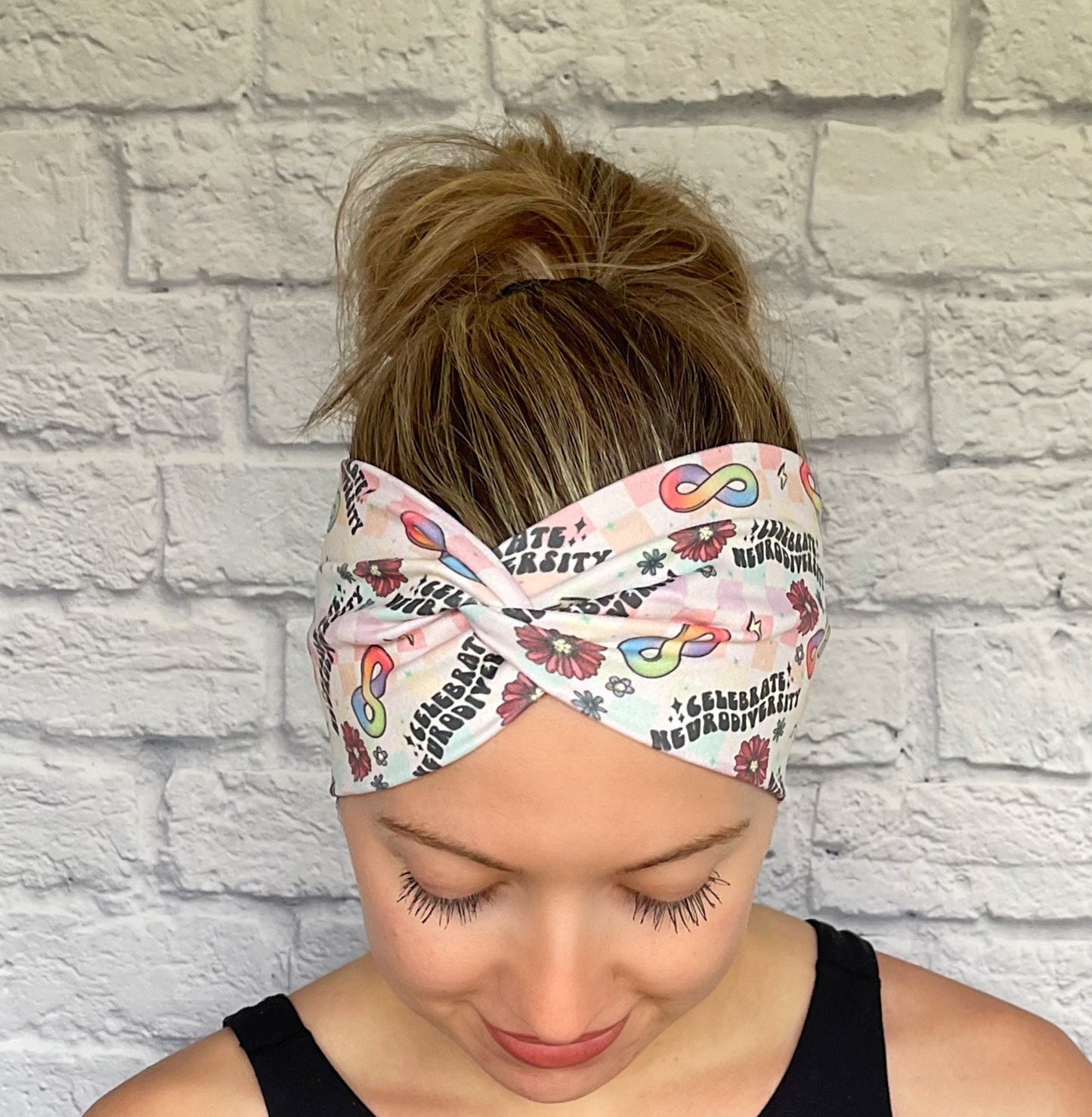 Woman with hair in bun wearing wide, twisted headband in rainbow plaid print with "celebrate neurodiversity", rainbow infinity signs, flowers, and lightning bolt print.