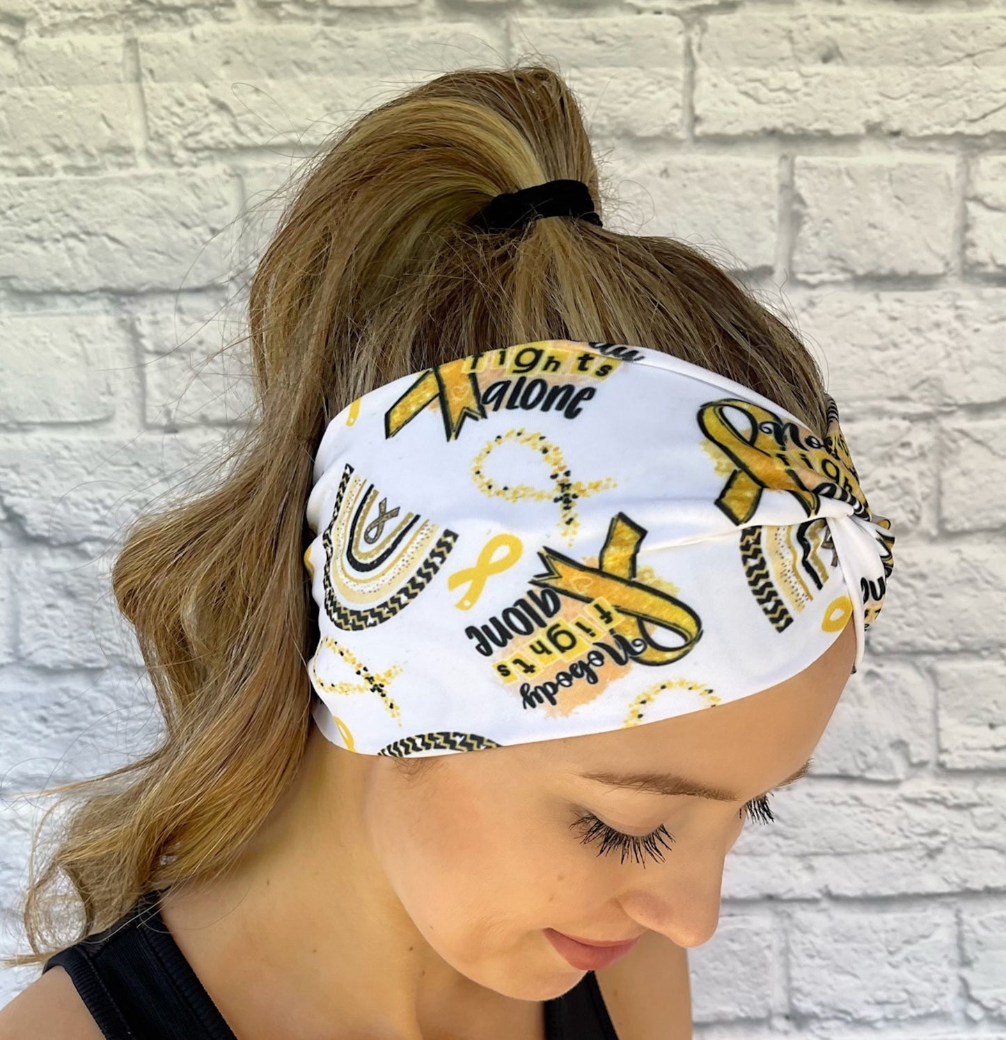 Woman with hair in ponytail wearing wide, twisted, white headband with "no one fights alone", gold ribbons, and gold/black rainbow print.