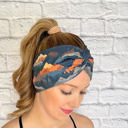 Woman with hair in curled ponytail wearing wide, twisted headband with mountain print in varying shades of blue, brown, and gray.