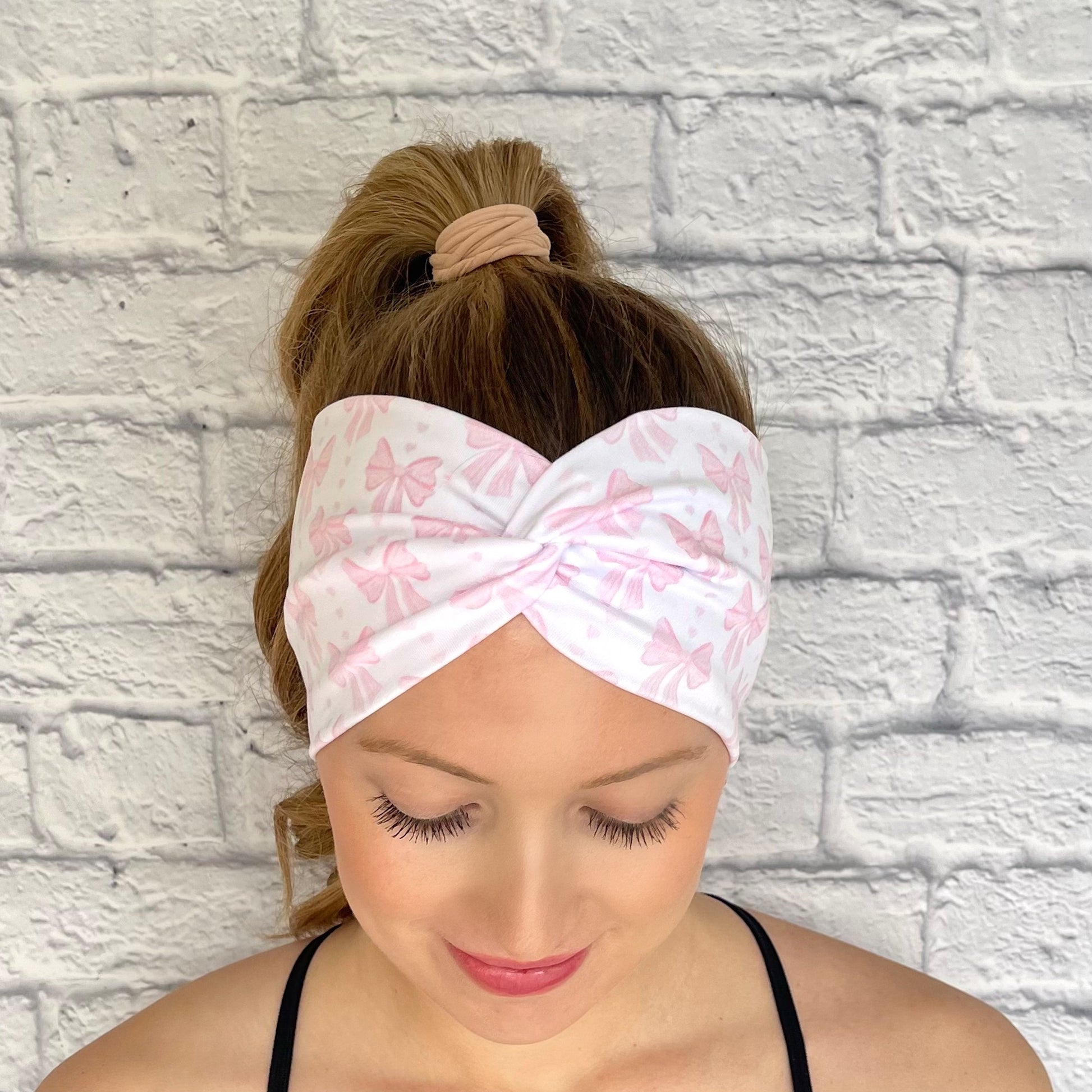 Woman with hair in curled ponytail wearing wide, twisted headband in white with light pink bow print.