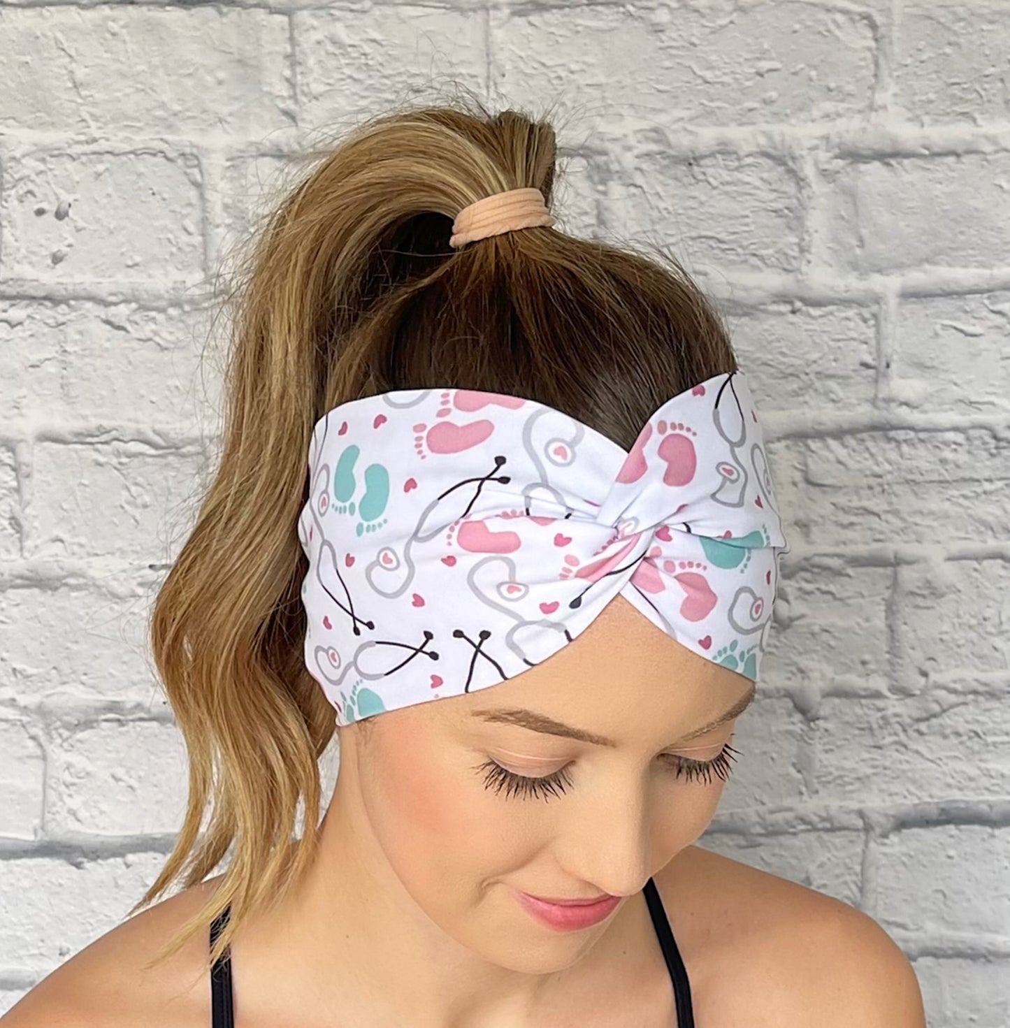 Woman wearing a white, OBGYN baby themed headband against a white brick wall.