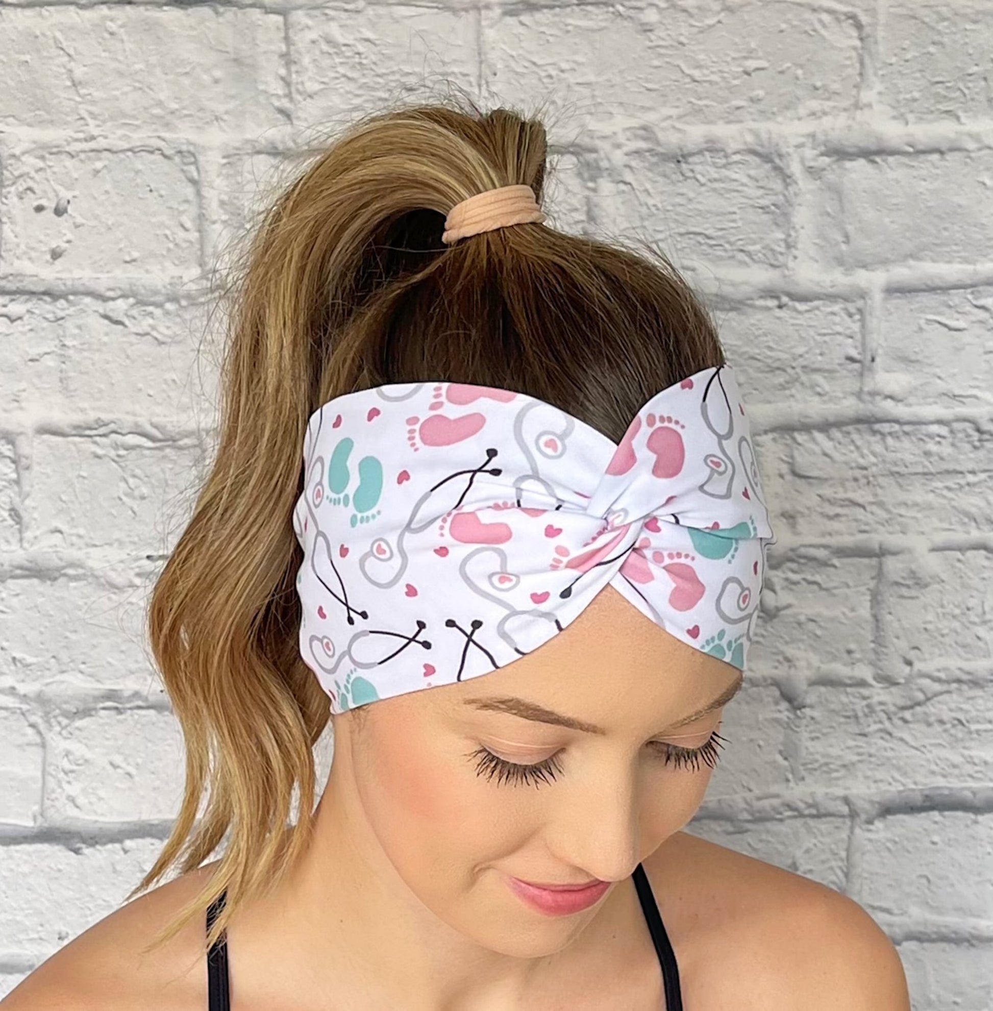 Woman wearing a white, OBGYN baby themed headband against a white brick wall.
