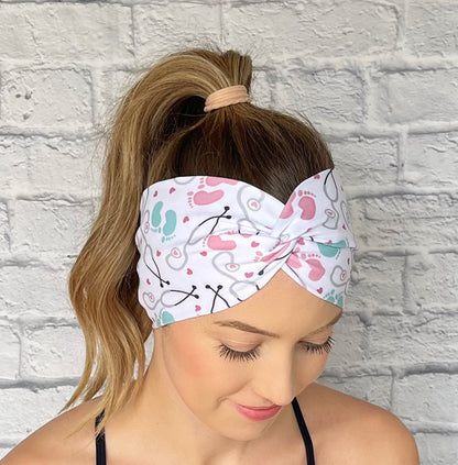 Woman wearing a white, OBGYN baby themed headband against a white brick wall.