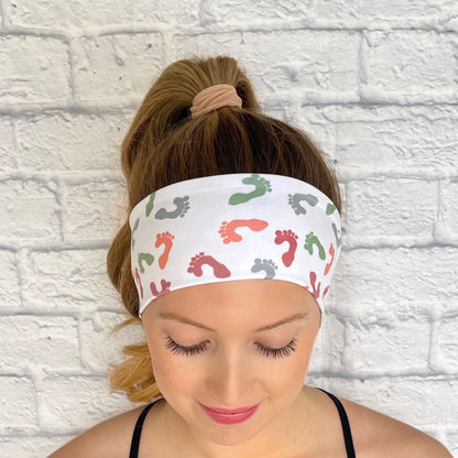 Woman with hair in curled ponytail wearing white headband with baby feet print in colors peach, wine, green, and light blue.