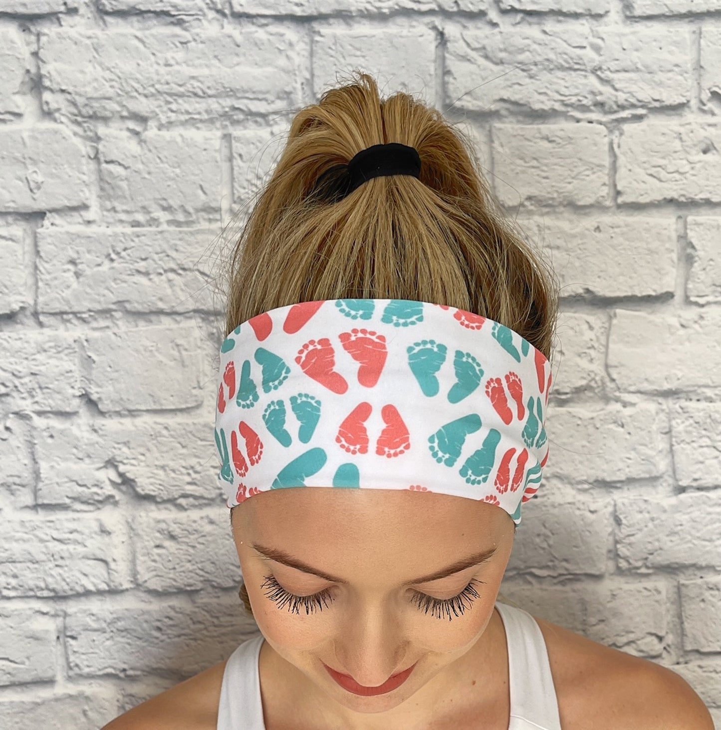 Woman with hair in ponytail wearing white, stretchy headband with pink and blue baby feet print.