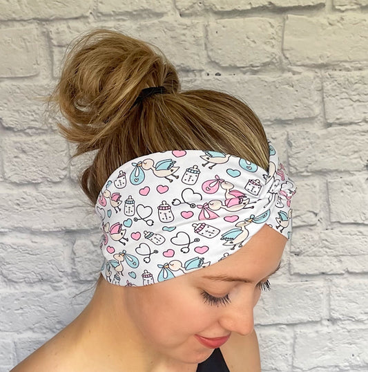 Woman with hair in bun wearing stretchy, wide, twisted headband in white with baby theme print- storks, hearts, bottles, and stethoscopes.