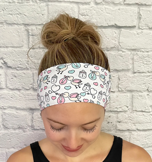 Woman with hair in bun wearing white, stretchy headband with baby theme print- storks, hearts, bottles, and stethoscopes.