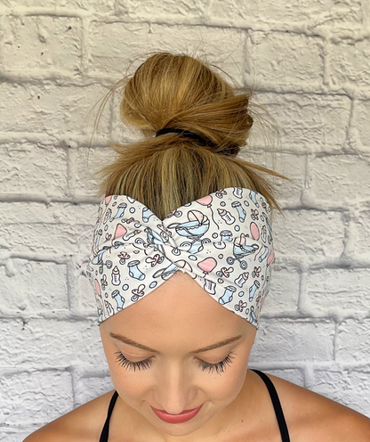 Woman with hair in bun wearing wide, twisted, cream headband with baby theme print- binkies, buggy, socks, bottles, and balloons.