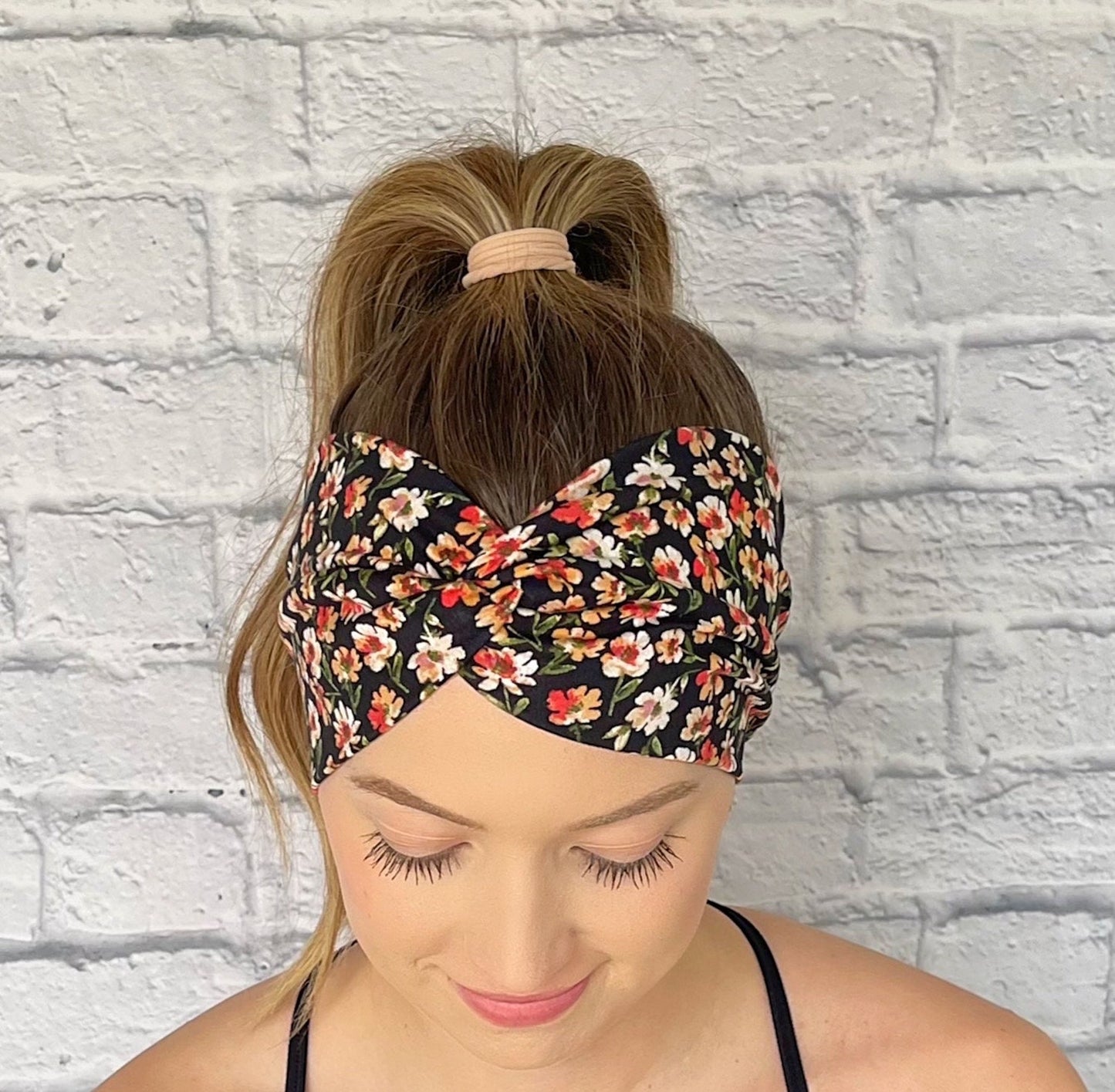 Woman with hair in curled ponytail wearing black wide twist headband with red, yellow, and white flower print.