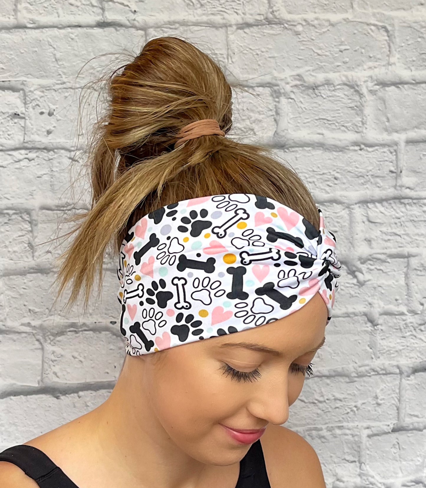 Woman with hair in bun wearing wide, twisted headband in white with black, pink, yellow, and light blue dog theme print with paws, dog bones, and hearts.