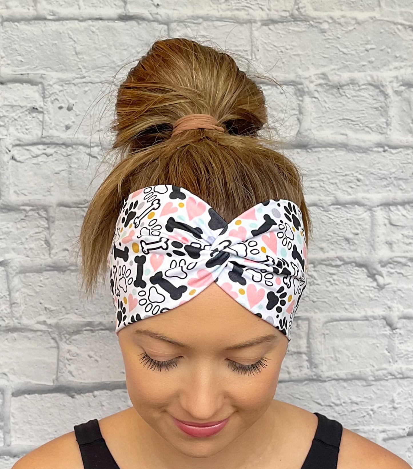 Woman with hair in bun wearing wide, twisted headband in white with black, pink, yellow, and light blue dog theme print with paws, dog bones, and hearts.