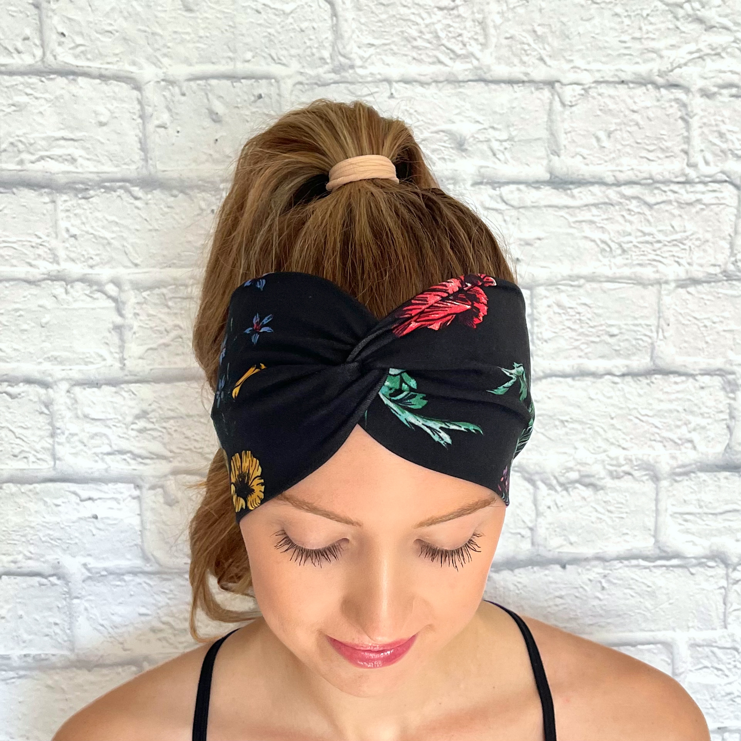 Woman with hair in curled ponytail wearing wide, twisted, black headband with red, yellow, blue, and green floral print.