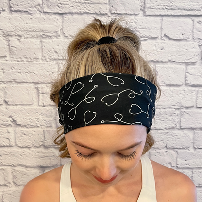 Woman with hair in curled ponytail with white brick background, wearing a black, stretchy headband with white stethoscope print.