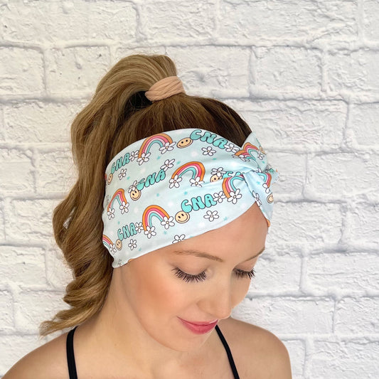 Woman with hair in curled ponytail wearing wide, twisted, light blue headband with "cna" print, rainbows, white flowers, and smiley faces.