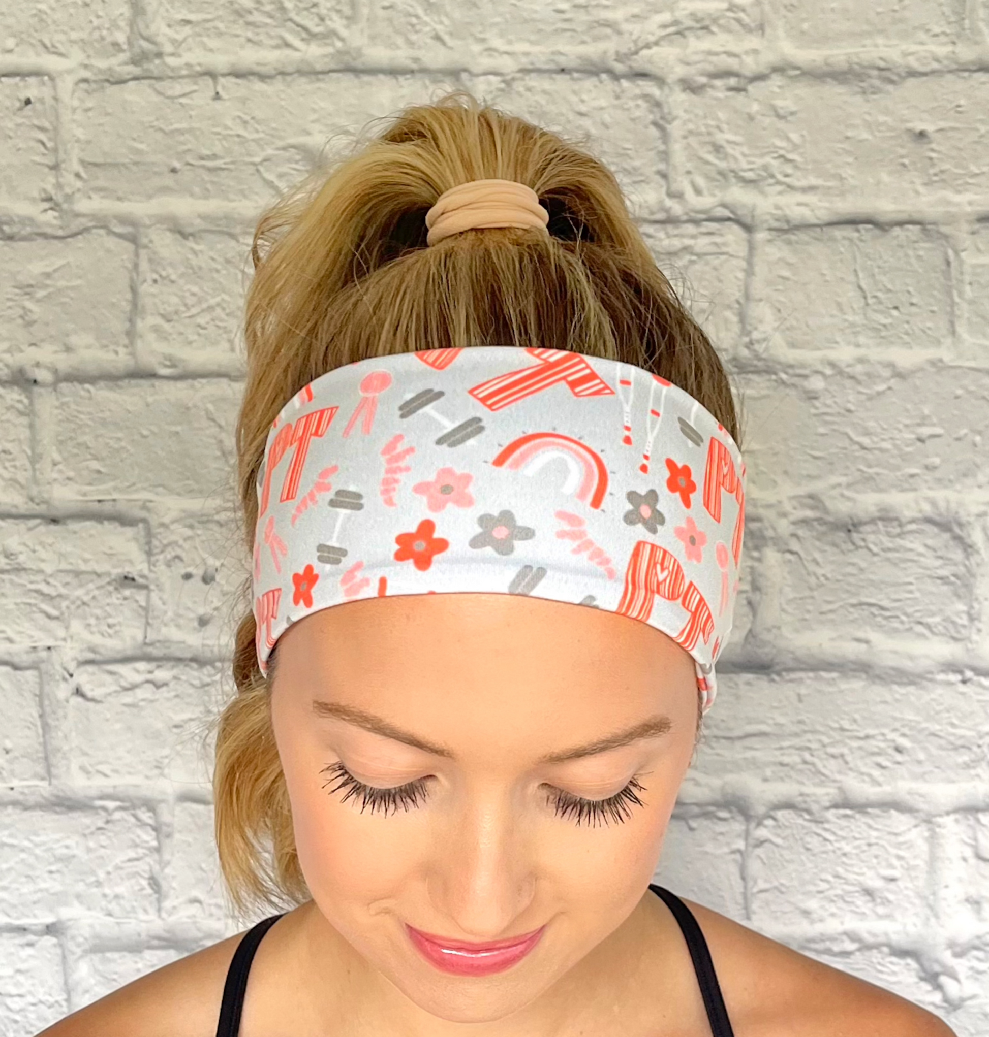 Woman with hair in curled ponytail wearing light blue/gray headband with "PT" print in orange and PT theme print with flowers in dark gray/blue and orange.