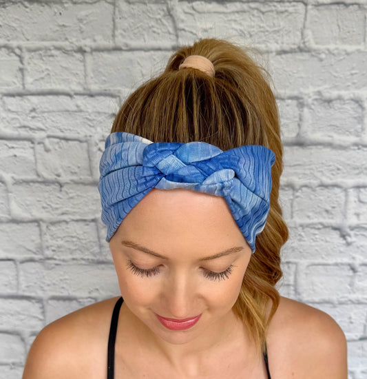 Woman with hair in curled ponytail wearing blue, ribbed, stretchy headband in sailor knot style with tie dye pattern.