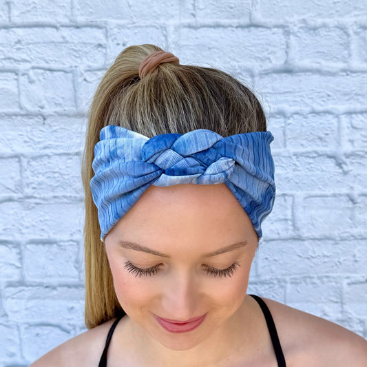 Woman with hair in ponytail wearing blue, ribbed, stretchy headband in sailor knot style with tie dye pattern.