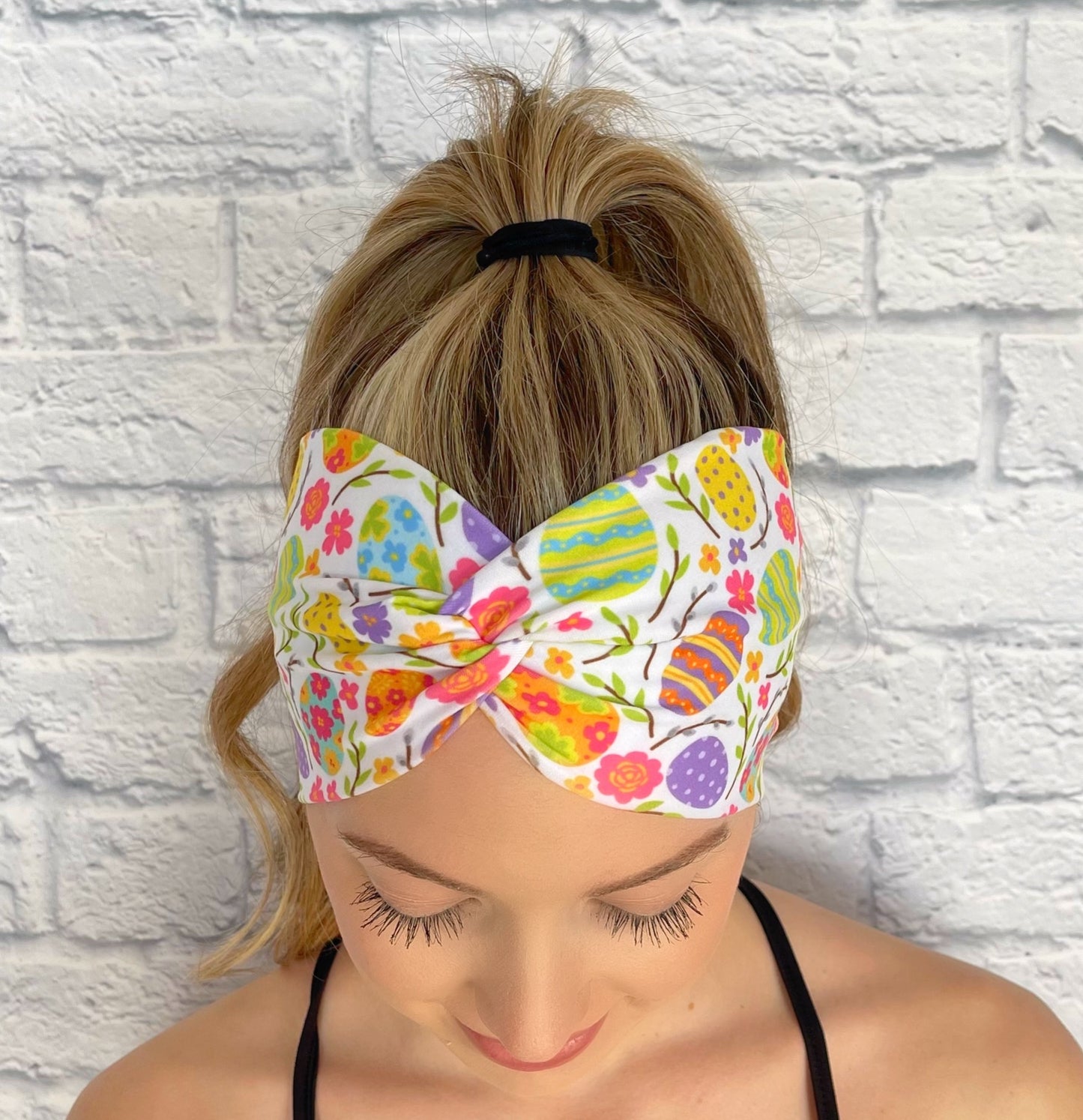 Woman with hair in curled ponytail wearing wide, twisted headband in white with bright color Easter egg print.