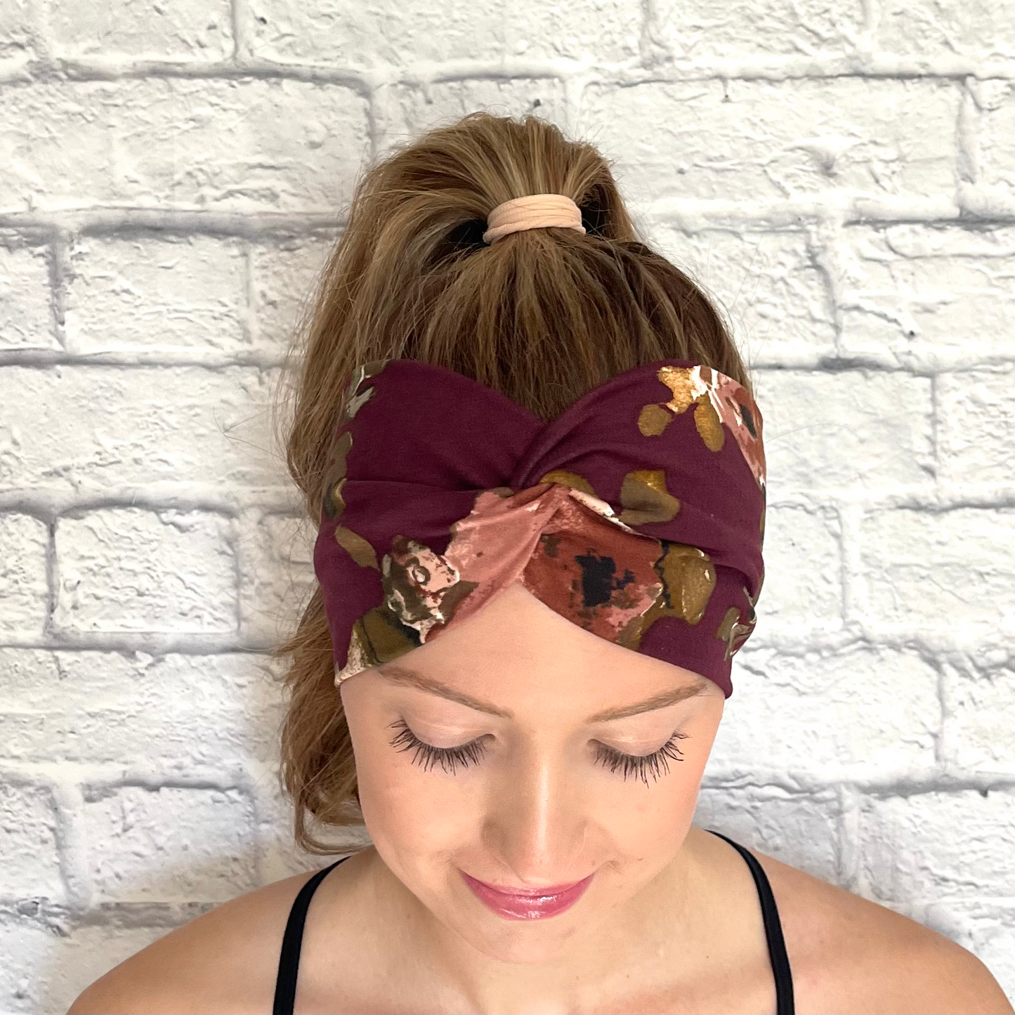 Woman with hair in curled ponytail wearing wide, twisted headband in maroon color with pink and gold flowers.