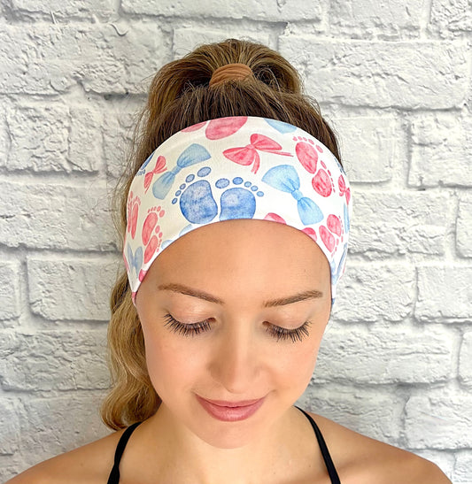 Woman with hair in ponytail wearing flat, stretchy, white headband with pink and blue baby feet and bow print.