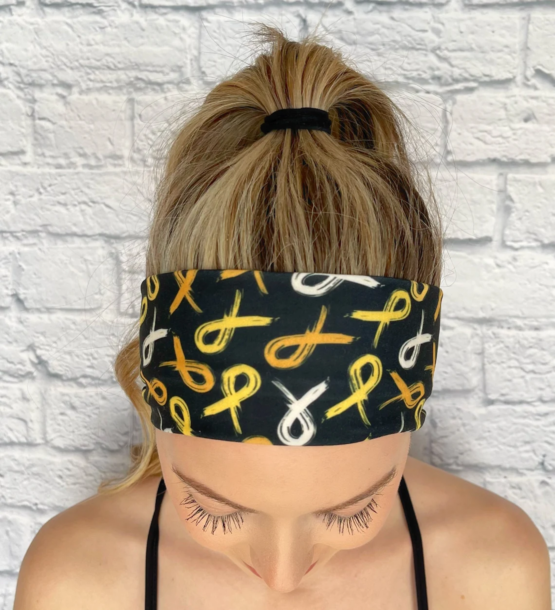 Woman with hair ini ponytail wearing flat, stretchy, black headband with gold ribbon print.