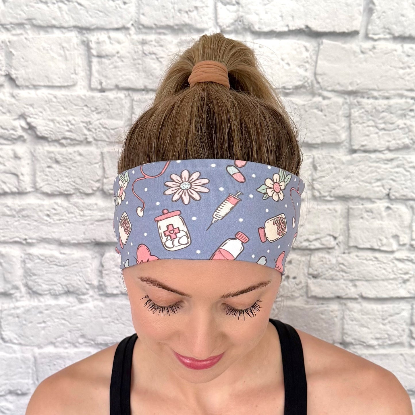 Woman with hair in ponytail wearing flat, stretchy headband in blue with pink, white, and mint color medical theme print.
