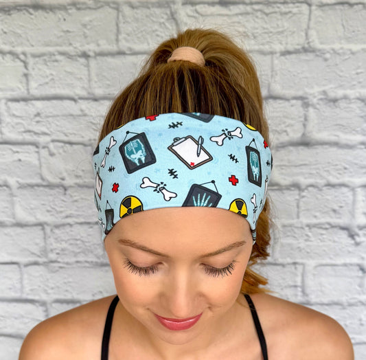 Woman with hair in ponytail wearing flat, stretchy, blue headband with radiology theme print displayed on a cream background.