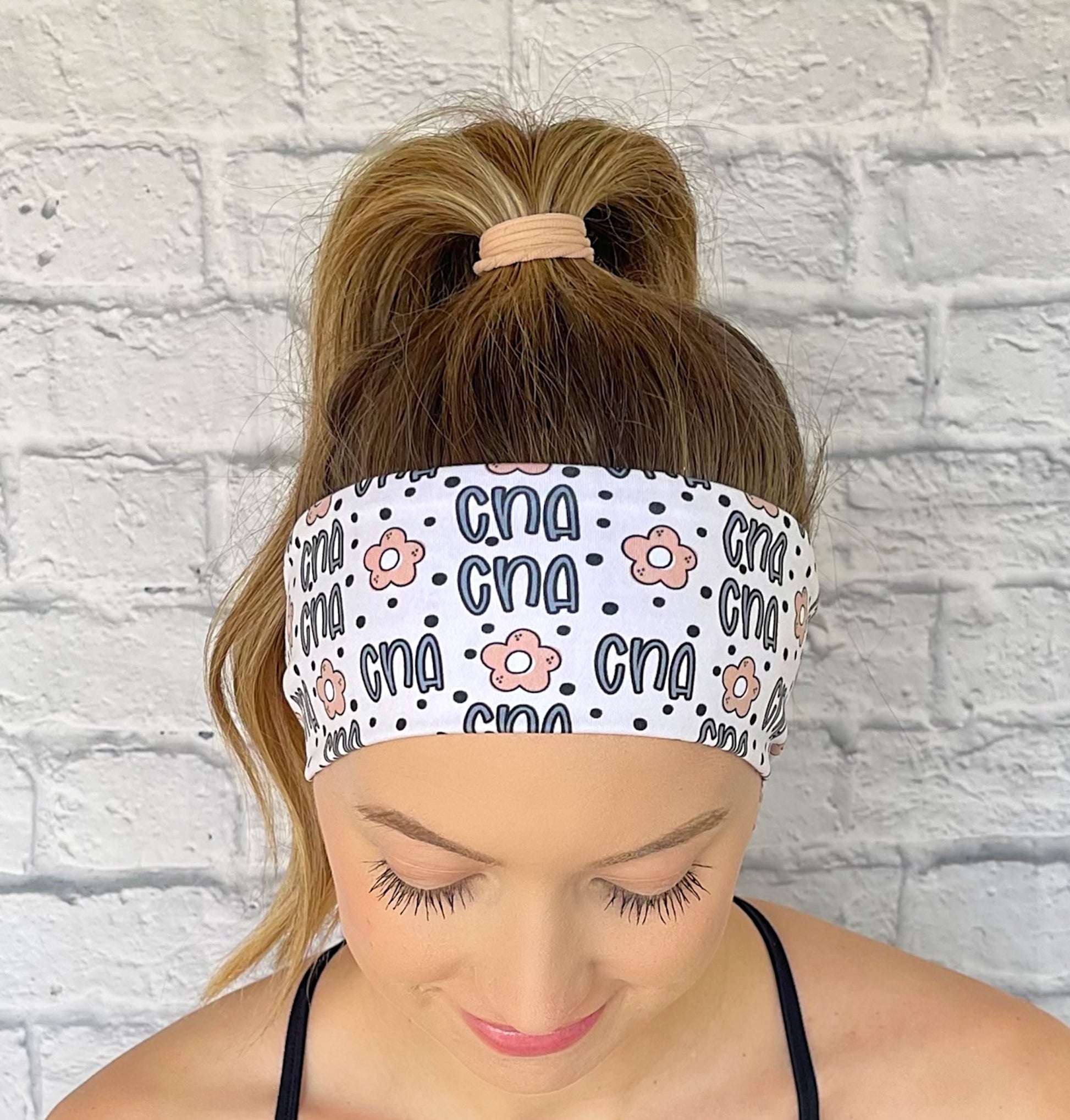 Woman with hair in curled ponytail wearing flat, stretchy, white headband with "cna", flowers, and polka dot print in blue and tan.