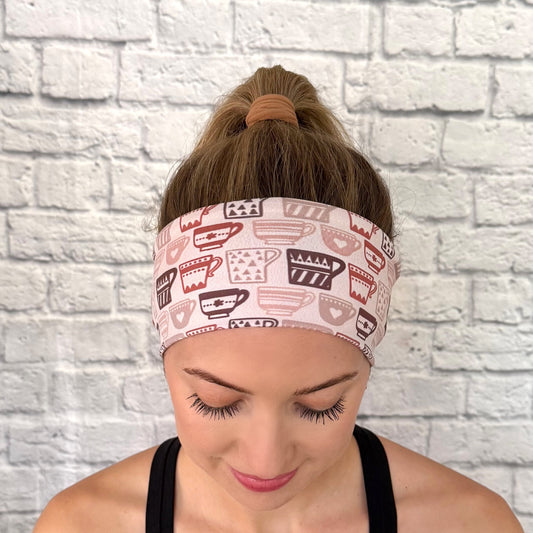 Woman wearing a headband with coffee cup pattern against a white brick wall.