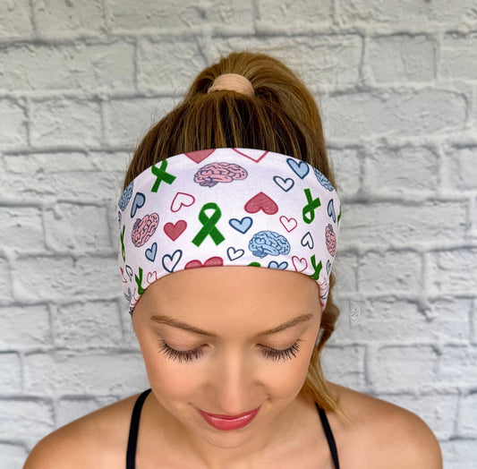 Woman with hair in ponytail wearing flat, stretchy headband with green ribbon, pink and blue brain, and pink and blue heart print.