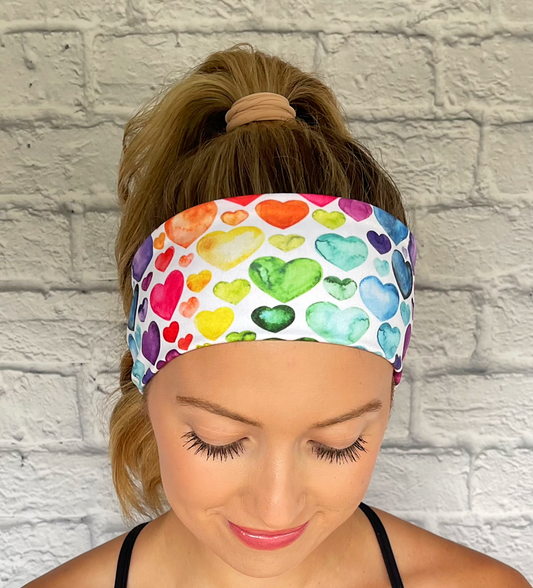 Woman with hair in curled ponytail wearing flat, stretchy headband in white with rainbow heart print.