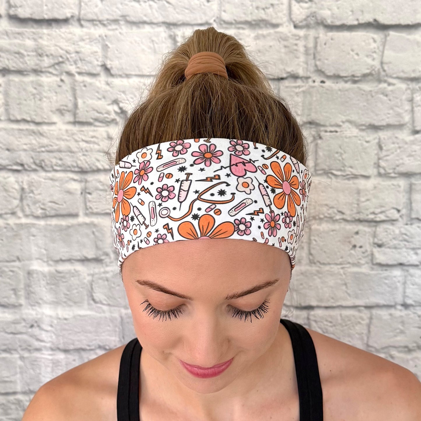 Woman wearing white headband with orange and pink medical theme print. Contains hearts, flowers, stethoscopes, bandages, and syringes.