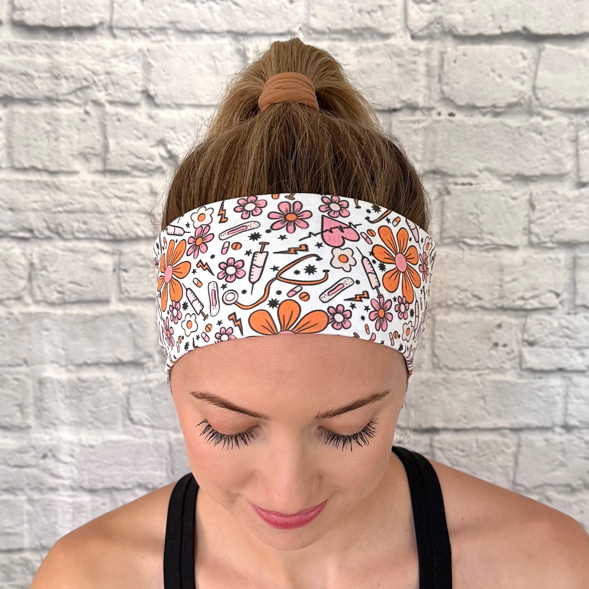 Woman wearing white headband with orange and pink medical theme print. Contains hearts, flowers, stethoscopes, bandages, and syringes.