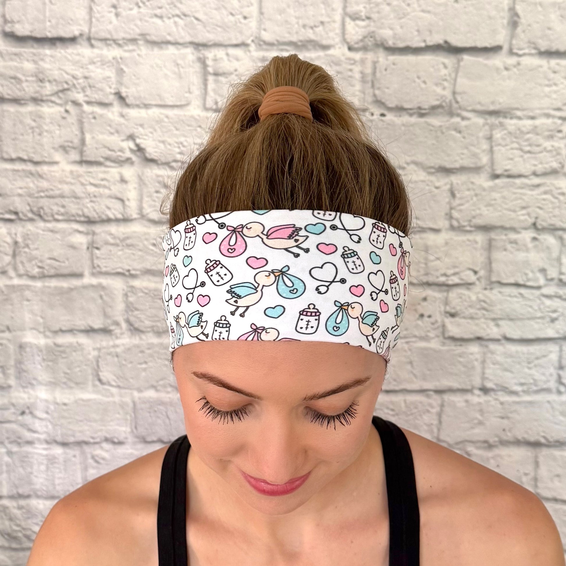Woman with hair in bun wearing white, stretchy headband with baby theme print- storks, hearts, bottles, and stethoscopes.