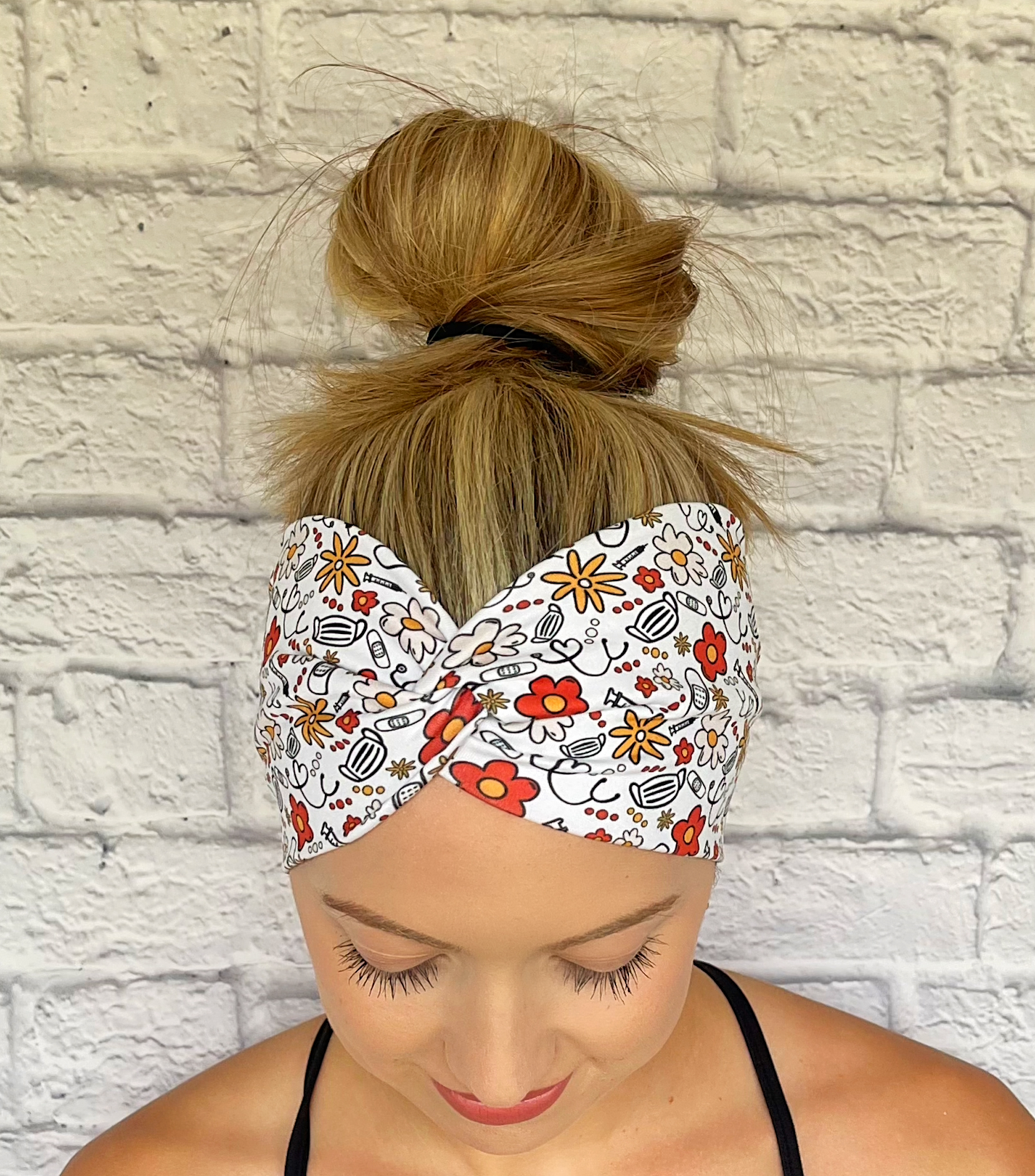 Woman with hair in bun wearing wide, twisted headband in white with red, yellow, and black floral and medical theme print.