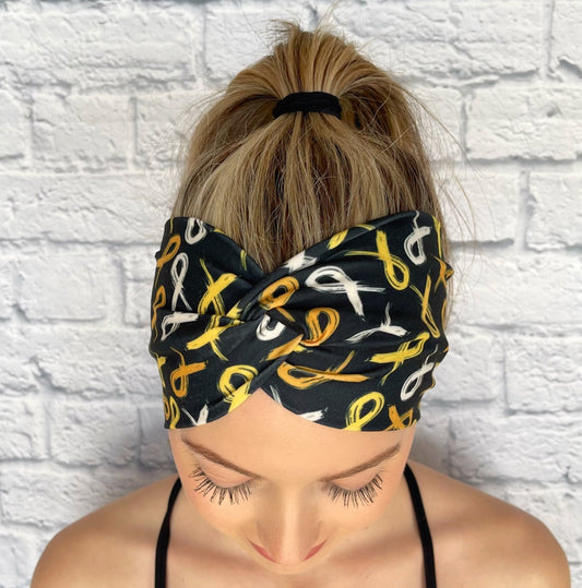 Woman with hair in ponytail wearing black, wide, twisted headband with gold ribbon print.