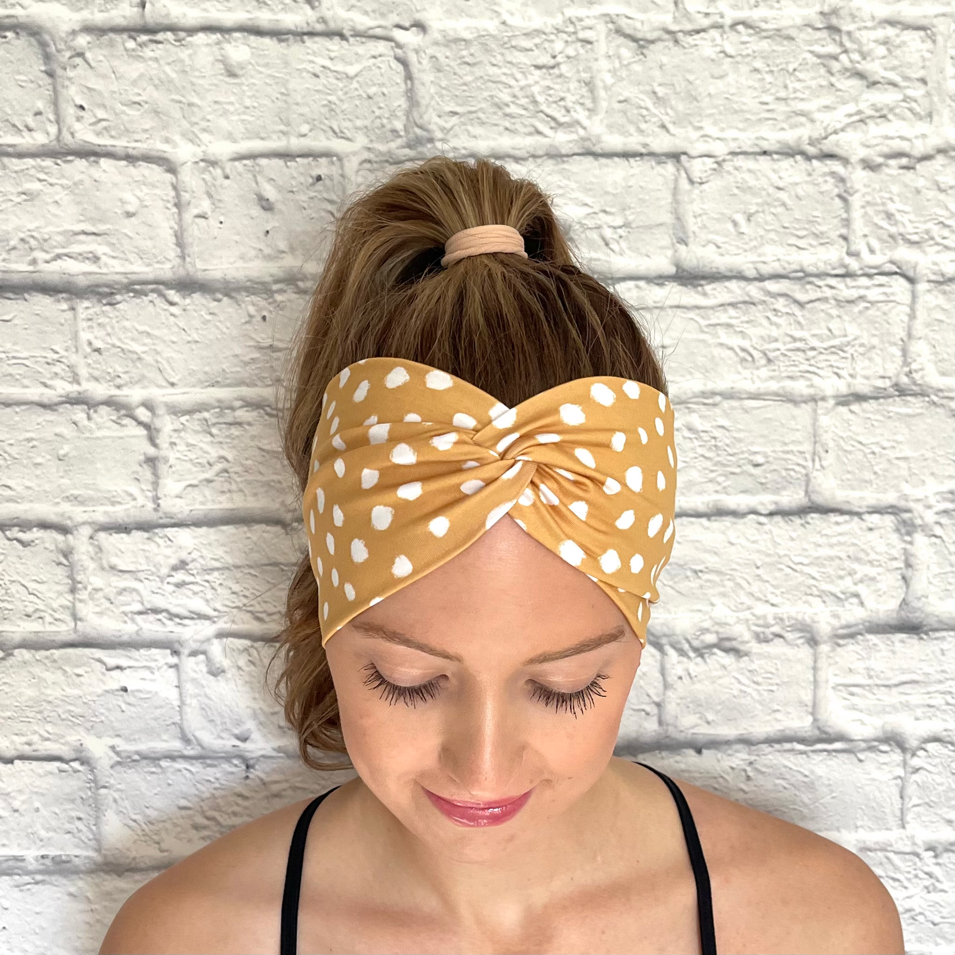 Woman with hair in curled ponytail wearing wide, twisted headband in gold color with white spots.