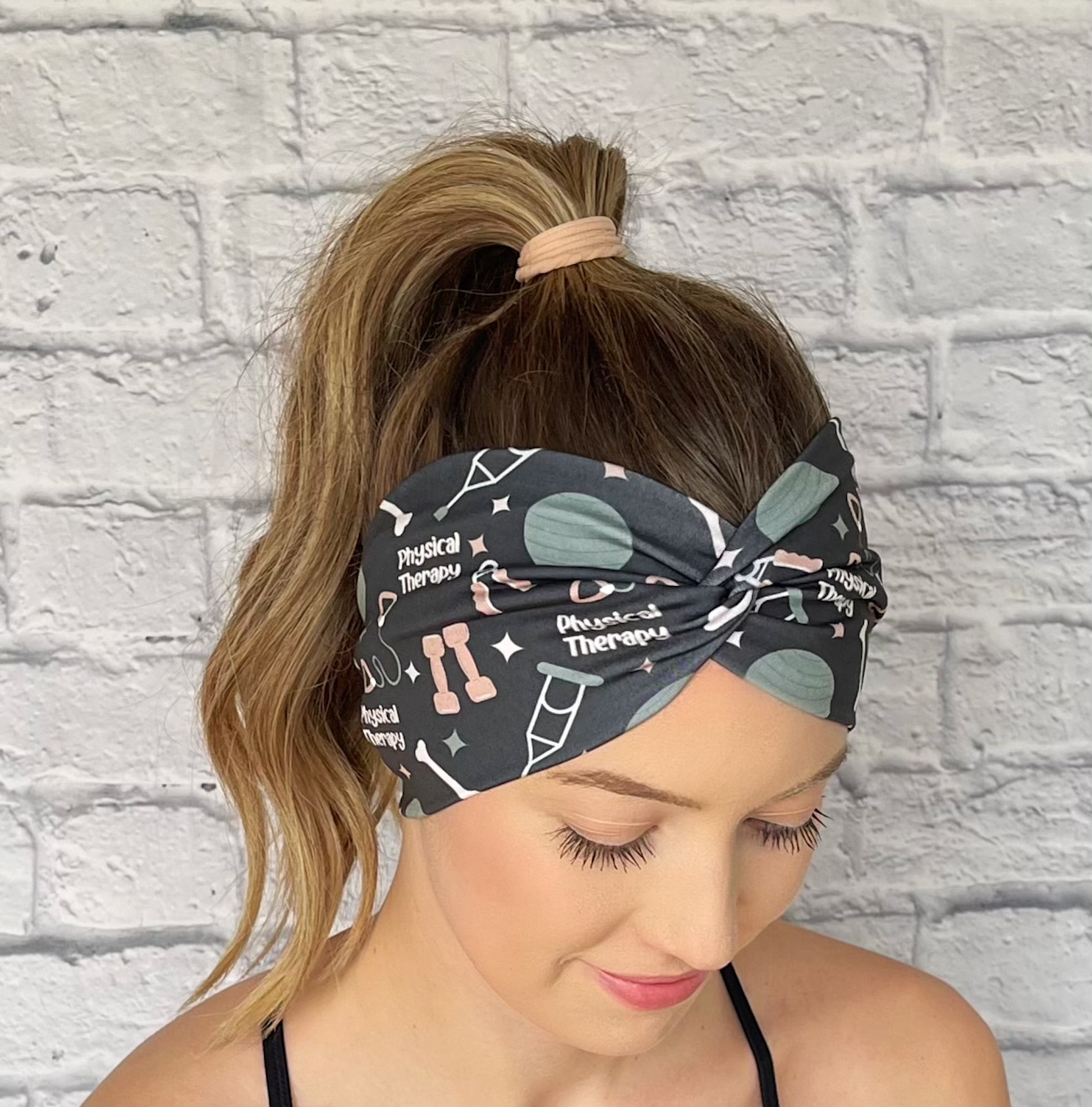 Woman with hair in curled ponytail wearing wide, twisted, gray/black headband with print "physical therapy" and exercise/therapy related items.