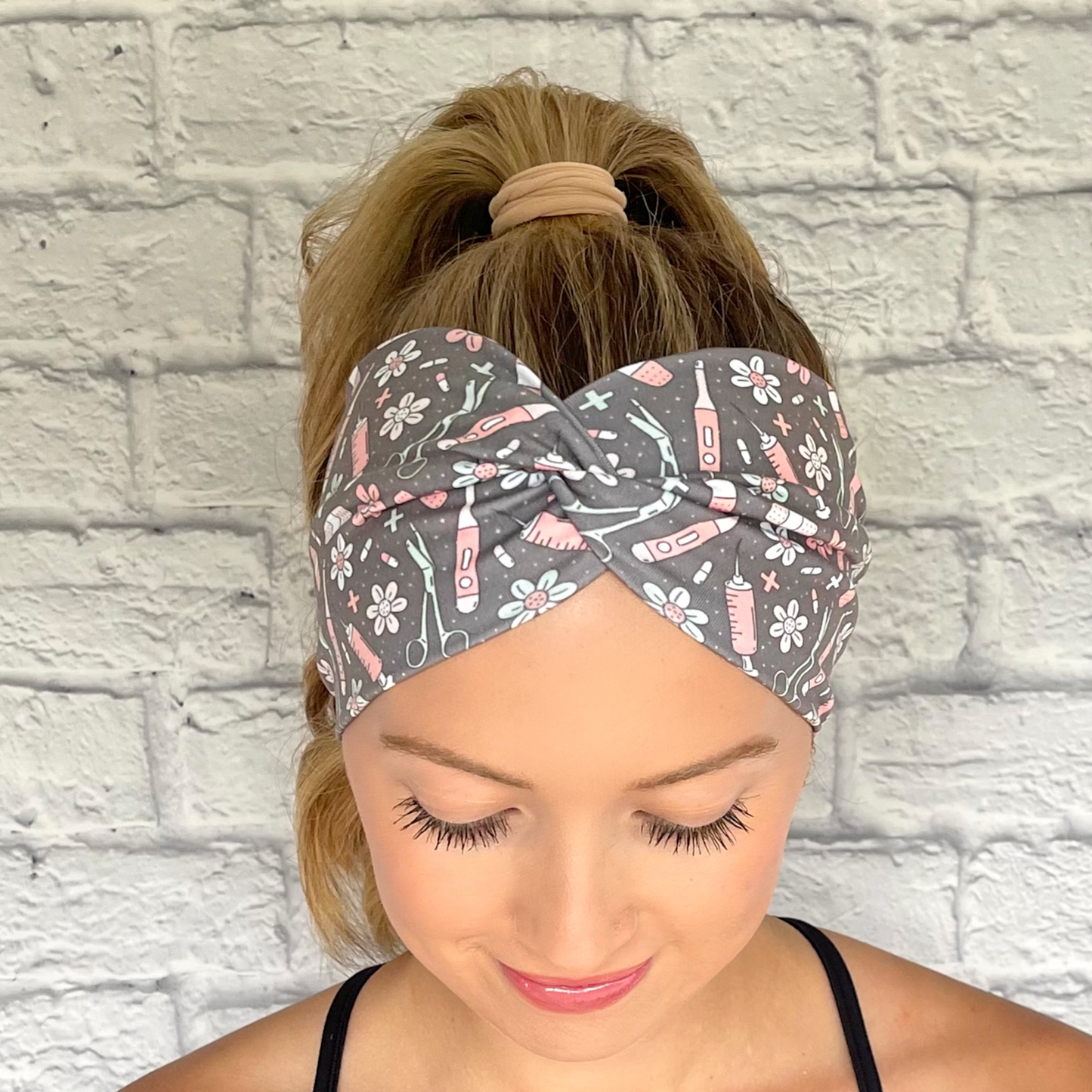 Woman with hair in curled ponytail wearing gray, wide, twisted headband with pink, mint blue, and white medical print.